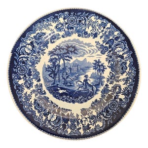 May include: A round, blue and white decorative plate with a detailed landscape scene in the center. The scene depicts a person on horseback, a dog, trees, and a building. The plate's border features floral patterns.