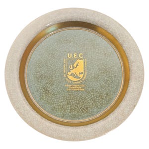 May include: A round decorative plate with a light green center and a gold rim. The center features the text "U.E.C" and a map of Europe. The outer edge is a textured off-white color. The plate has text that reads "8.ème Congrès UEC Copenhague 7-10 octobre 1969."