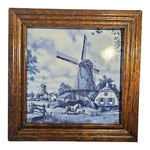 Può includere: Opera d'arte quadrata incorniciata con una scena di mulino a vento olandese blu e bianca. L'opera raffigura mulini a vento, case e un cavallo in un campo. La cornice è in legno con finitura naturale.