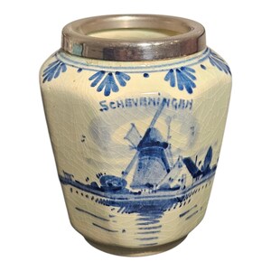 Könnte beinhalten: Ein Vintage-Keramiktopf mit silbernem Rand, der eine blau-weiße Windmühlenszene und das Wort "Scheveningen" zeigt. Der Topf hat ein leicht rissiges, gealtertes Aussehen, was seinen antiken Charme unterstreicht.