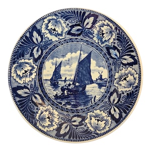 May include: A round, blue and white decorative plate. The center features a nautical scene with sailboats and a windmill. The border is adorned with floral and leaf patterns. The plate has a glossy finish.