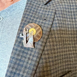 May include: A copper-toned pin featuring a howling wolf silhouette against a golden sun and a cactus. The pin is attached to a gray sweater and a blue and gray plaid blazer. The pin's design evokes a desert landscape.