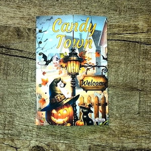 Könnte beinhalten: Ein Halloween-Schild mit den Worten "Candy Town" über einer beleuchteten Straßenlaterne und einem geschnitzten Kürbis mit Hexenhut. Eine schwarze Katze mit Hexenhut sitzt in der Nähe eines Holzzauns mit einem "Welcome"-Schild.