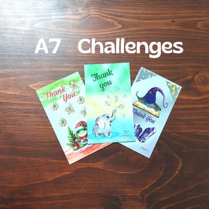 Könnte beinhalten: Drei rechteckige Karten mit der Aufschrift A7 Challenges. Die linke Karte zeigt einen Frosch mit Weihnachtsmütze und Lebkuchen. Die mittlere Karte zeigt einen Elefanten, der Blasen bläst, mit dem Text Thank you. Die rechte Karte zeigt einen Zauberhut und Kristalle.