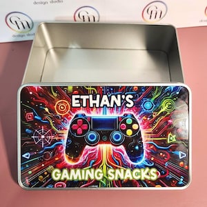 May include: A rectangular metal tin with an open lid. The lid displays a colourful design featuring a video game controller, the name "ETHAN'S", and the words "GAMING SNACKS". The background is a vibrant mix of red, blue, yellow, and green.