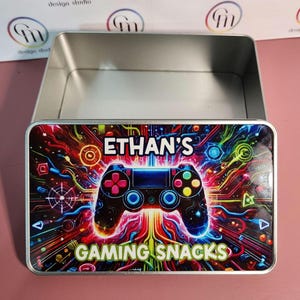 May include: A rectangular metal tin with a hinged lid. The lid features a colourful design with a video game controller and the text "ETHAN'S GAMING SNACKS". The background is a vibrant mix of red, blue, and yellow.