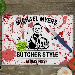 May include: A rectangular cutting board with a white background featuring a graphic of Michael Myers, a knife, and red splatter designs. Text includes "MICHAEL MYERS," "BUTCHER STYLE," and "ALWAYS FRESH."