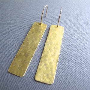 May include: A pair of gold-colored, rectangular earrings with a hammered texture. The earrings hang from thin, curved ear wires. The earrings are displayed against a gray background.