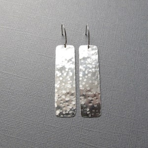 Puede incluir: Un par de pendientes colgantes rectangulares plateados con textura martillada. Los pendientes tienen un diseño de gancho simple y un acabado pulido. Los pendientes se muestran sobre un fondo gris.