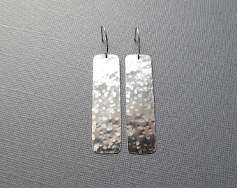 May include: A pair of silver rectangular dangle earrings with a hammered texture. The earrings have a simple hook design and a polished finish. The earrings are displayed against a gray background.
