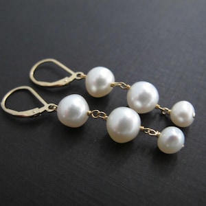 May include: A pair of elegant drop earrings with white pearls. Each earring has a gold-coloured leverback clasp and is strung with three pearls of varying sizes, connected by delicate gold-toned chains.