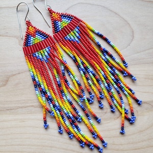 May include: A pair of long, dangling earrings crafted from small, multi-colored glass beads in shades of red, orange, yellow, green, and blue. The earrings feature a triangular top section with a geometric pattern and long, flowing beaded fringe.