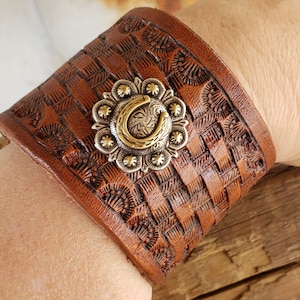 May include: Brown leather cuff bracelet with a silver and gold horseshoe design. The bracelet has a woven pattern and is decorated with a floral design.