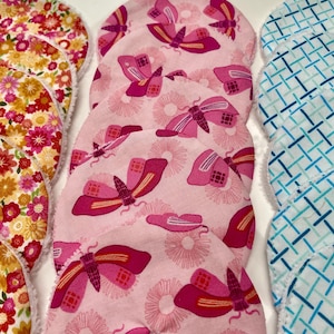 May include: Several circular fabric items with diverse patterns. Some display pink butterflies on a pink backdrop, others have floral designs in red, yellow, and pink, and some feature a blue and white geometric pattern.