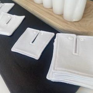 May include: A stack of white cotton fabric squares with a buttonhole in the centre. The squares are sewn with white thread and have a white border.