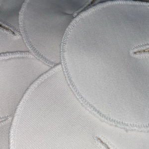 May include: Close-up of several white, circular fabric pads with a stitched edge and a small slit. One pad has a snap closure. The pads are stacked and slightly overlapping, showing the texture of the fabric.