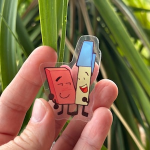 May include: A clear sticker featuring cartoon illustrations of a red eraser and a blue pen, both with smiling faces and holding hands. The background is blurred green foliage.