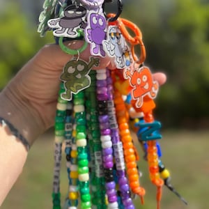 May include: A collection of colorful keychains featuring cartoon character designs. Each keychain has a beaded lanyard with various colors and the words "FOURTEEN", "SIX", "FIVE", and "BFF" spelled out. The keychains are attached to a variety of colored rings.