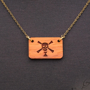 May include: A gold chain necklace with a wooden pendant featuring a skull and crossbones design. The pendant is rectangular and has a reddish-brown color.