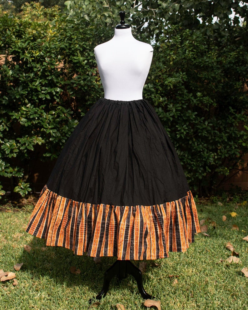 Op de afbeelding: Zwarte rok met een brede, gerimpelde zoom met verticale oranje en zwarte strepen. De rok wordt getoond op een witte mannequin tegen een achtergrond van groen gebladerte. De rok is een volle, vloeiende stijl.