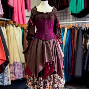 May include: A brown and purple Renaissance-style dress on a mannequin. The dress features a dark purple corset, a layered skirt, and long sleeves with pink ribbons. The bottom layer of the skirt has a skull and floral print.