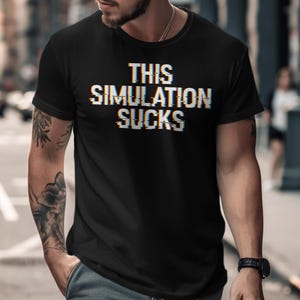Puede incluir: Camiseta negra de cuello redondo con el texto "THIS SIMULATION SUCKS" en efecto glitch. La camiseta la lleva una persona con tatuajes en el brazo. El fondo es una calle de la ciudad borrosa.