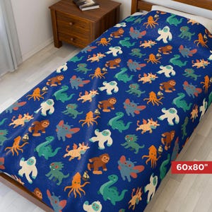 May include: A blue blanket with a repeating pattern of colorful cartoon monsters. The blanket measures 60x80 inches. The monsters are in various colors, including orange, green, brown, and white. The blanket is on a bed with a wooden frame.