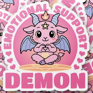 May include: Pink sticker featuring a cartoon demon with blue wings, horns, and a flame. The sticker has the words "EMOTIONAL SUPPORT DEMON" in white and pink. Hearts and a pentagram are also included in the design.