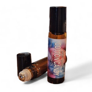 May include: Two brown glass roll-on perfume bottles with black caps. One bottle is upright, the other is lying down with the cap off. The label reads "Cotton Candy Perfume Oil" with a colorful background.