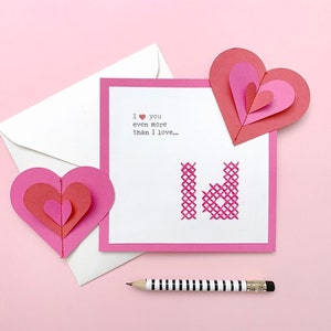 Può includere: Un biglietto rosa con un cuore rosa cucito su di esso. Il biglietto recita "Ti amo ancora di più di quanto amo..." e ha una busta rosa accanto. Ci sono due cuori di carta rosa sul biglietto.