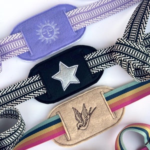 Puede incluir: Tres cintas de tela diferentes con diseños bordados. Las cintas son de color negro y blanco, arcoíris y morado con blanco. Los diseños son una estrella, un pájaro y un sol.