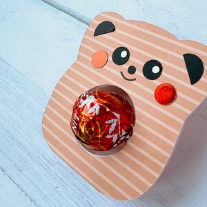 Puede incluir: Una cara de panda de cart&oacute;n a rayas rosa y blancas con orejas y ojos negros. El panda tiene un caramelo de chocolate rojo y dorado en la boca.