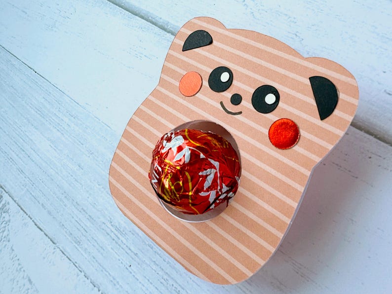 Puede incluir: Una cara de panda de cart&oacute;n a rayas rosa y blancas con orejas y ojos negros. El panda tiene un caramelo de chocolate rojo y dorado en la boca.