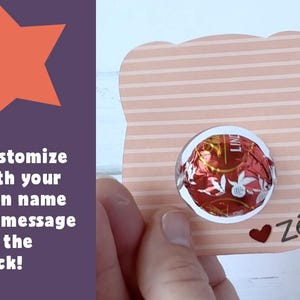 Puede incluir: Una tarjeta de rayas rosa claro y blanco con un recorte redondo para un chocolate. La tarjeta tiene un coraz&oacute;n rojo y el texto "Z&eacute;" escrito en negro. La parte posterior de la tarjeta se puede personalizar con un nombre o un mensaje.