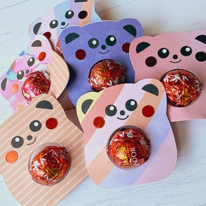 Puede incluir: Seis soportes de cart&oacute;n en forma de oso de diferentes colores y patrones, con un agujero en el centro para un caramelo. Los osos est&aacute;n sobre una superficie de madera blanca.