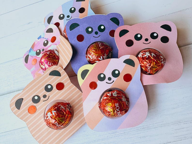 Puede incluir: Seis soportes de cart&oacute;n en forma de oso de diferentes colores y patrones, con un agujero en el centro para un caramelo. Los osos est&aacute;n sobre una superficie de madera blanca.