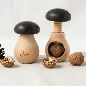 Op de afbeelding: Twee houten notenkrakers in de vorm van paddenstoelen. De grotere notenkraker heeft een zwarte bovenkant en de kleinere een bruine bovenkant. Beide notenkrakers hebben een houten basis en een schroefmechanisme om noten te kraken. De kleinere notenkraker heeft het woord "Anton" gegraveerd op de zijkant.