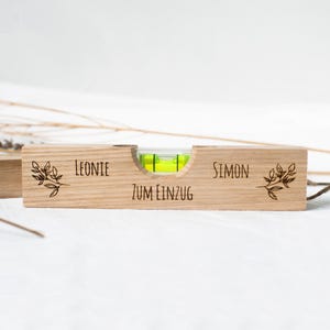 Op de afbeelding: Een houten waterpas met een groene waterpas en de woorden "Leonie" en "Simon" gegraveerd aan beide kanten. De waterpas heeft ook de woorden "Zum Einzug" gegraveerd in het midden.