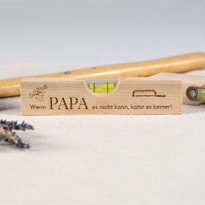 Pode incluir: Um nível de madeira com a palavra "PAPA" gravada, juntamente com a frase "Wenn es nicht kann, kann es keiner!". O nível tem um frasco transparente com um líquido amarelo e uma bolha. Uma chave inglesa e uma serra também estão gravadas.