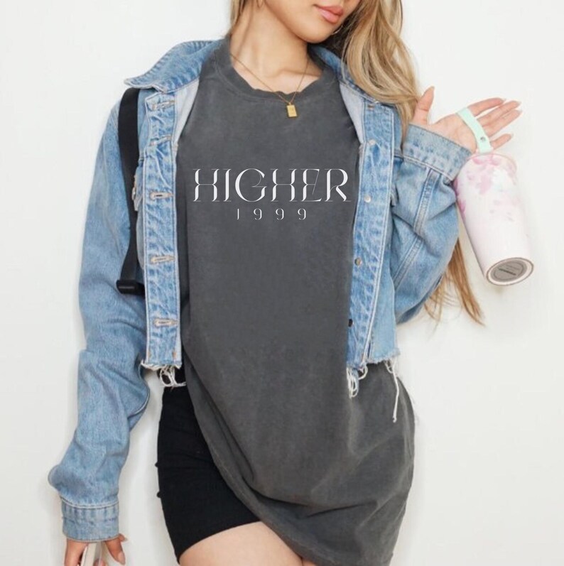 Peut inclure: Une femme portant un t-shirt gris fonc&eacute; avec le texte "HIGHER 1999" en blanc. Elle porte &eacute;galement une veste en jean bleu clair et un short noir.
