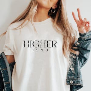 Peut inclure: Un t-shirt blanc avec le texte "HIGHER 1999" imprim&eacute; en noir.