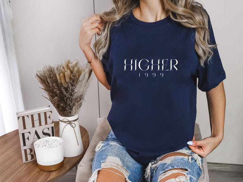 Peut inclure: Un t-shirt bleu marine avec le mot "HIGHER" en lettres blanches et l'ann&eacute;e "1999" en dessous. Le t-shirt est port&eacute; par une personne aux longs cheveux blonds.