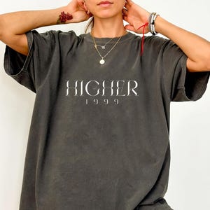 Peut inclure: Un t-shirt surdimensionn&eacute; gris fonc&eacute; avec le texte "HIGHER 1999" imprim&eacute; en blanc sur le devant.