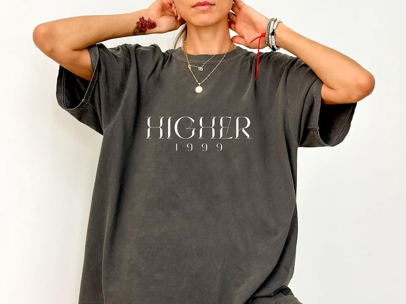 Peut inclure: Un t-shirt surdimensionn&eacute; gris fonc&eacute; avec le texte "HIGHER 1999" imprim&eacute; en blanc sur le devant.