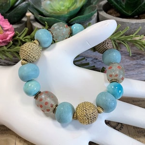 May include: A turquoise and gold beaded bracelet with a woven gold bead and speckled turquoise beads.