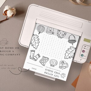 May include: A black and white printable Cozy Autumn word search puzzle sheet is shown inside a white printer. The sheet features fall-themed illustrations and the text "Print at home or through a printing company".
