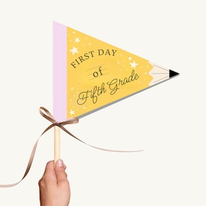 Op de afbeelding: Een gele wimpel met de tekst "FIRST DAY of Fifth Grade" en sterren. De wimpel is ontworpen als een potlood. Het is bevestigd aan een houten deuvel met een bruin lint dat in een strik is geknoopt.