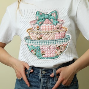 May include: White t-shirt with a graphic of three decorative bowls, each with a different floral or gingham pattern. A large bow sits on top. The text "Tangled Soul" is visible on the design.