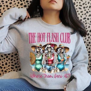 Op de afbeelding: Een lichtgrijs sweatshirt met een afbeelding van drie skeletfiguren in badpakken en zonnehoeden, die drankjes en een waaier vasthouden. De tekst "THE HOT FLASH CLUB" en "where them fans at?" staan boven en onder de afbeelding.