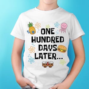 May include: White t-shirt featuring the text "ONE HUNDRED DAYS LATER..." with cartoon graphics of a pineapple, jellyfish, burger, and a character. The shirt has a crew neck and short sleeves.